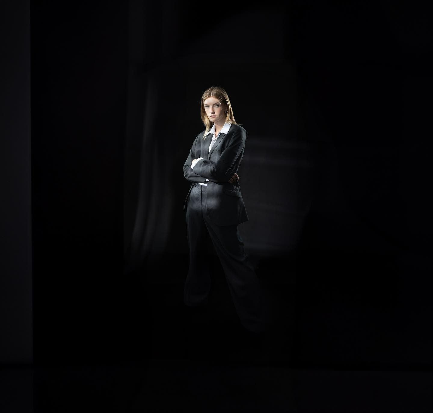 Woman in a dark suit stands with arms crossed in a dim room; light falls on her.