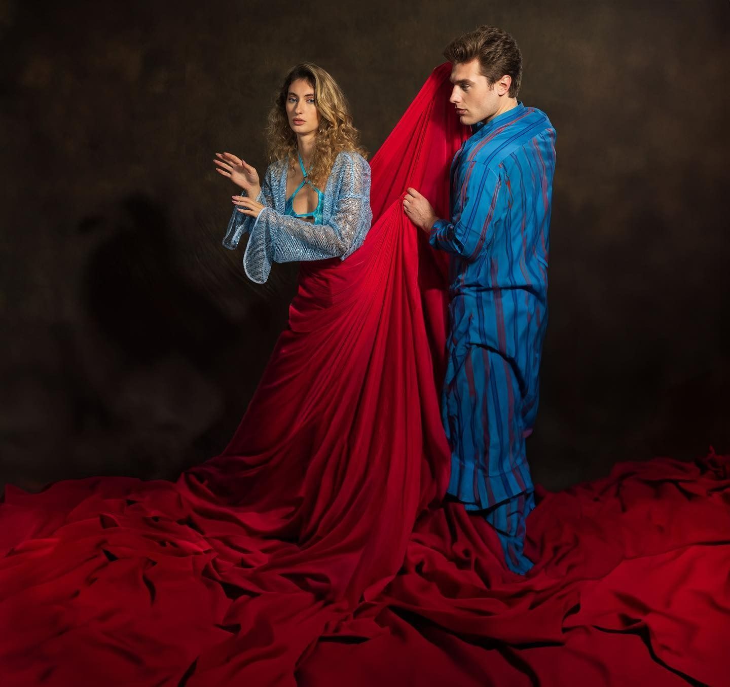 Woman in blue, man in blue, red fabric drape. Studio setting, woman gestures, man holds fabric.