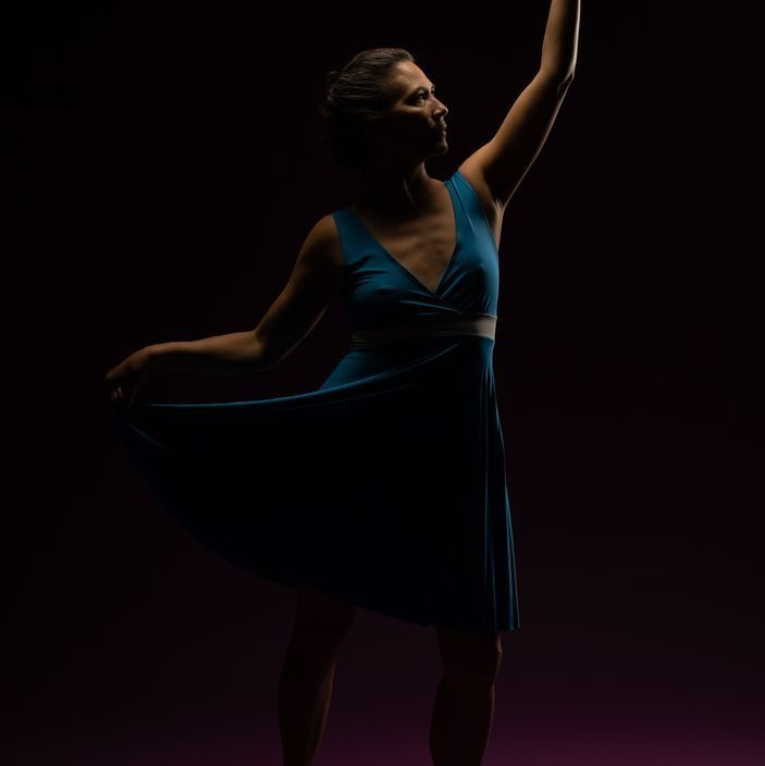 A dancer in a teal dress, arms raised, poised against a dark purple background.