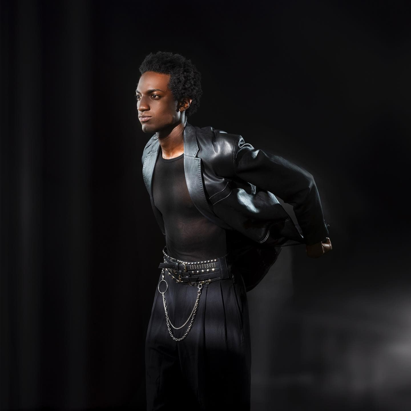 Man in black leather jacket, sheer shirt, and pleated pants, striking a pose. Dark background.