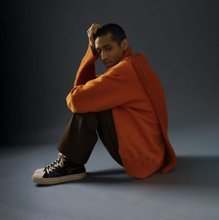 Man in orange sweater and brown pants sits on floor, hand on head, looking down. Black sneakers.