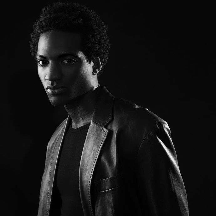 Man in a leather jacket, looking toward the viewer with a serious expression. Black and white, against a dark background.
