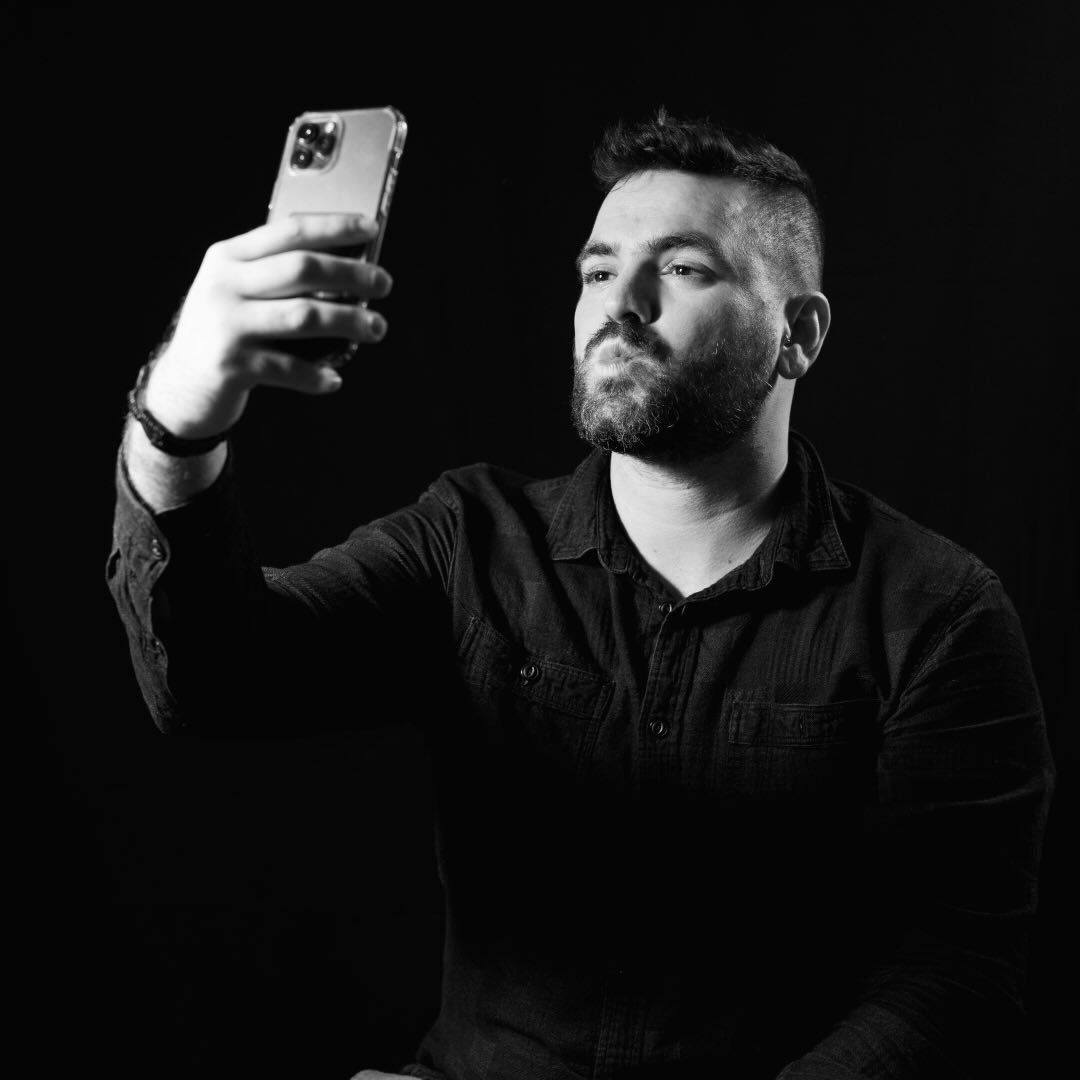 Man holding smartphone, taking a selfie. Black shirt, dark background.