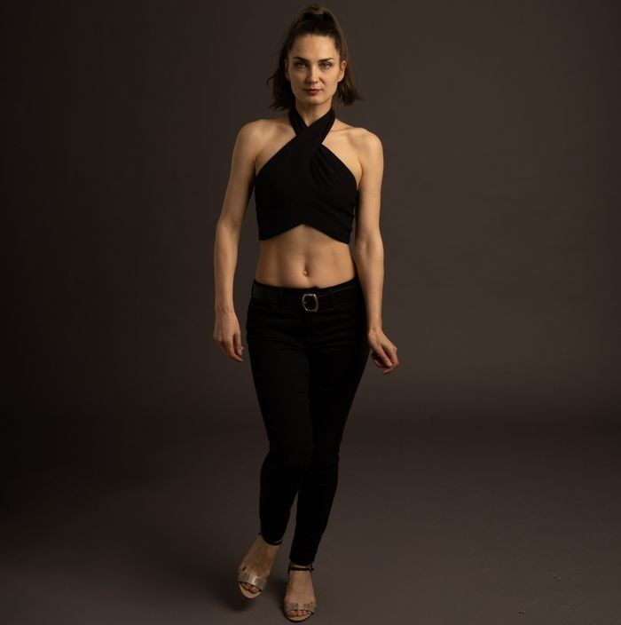 Woman in black halter top and pants, walking forward, gray background.