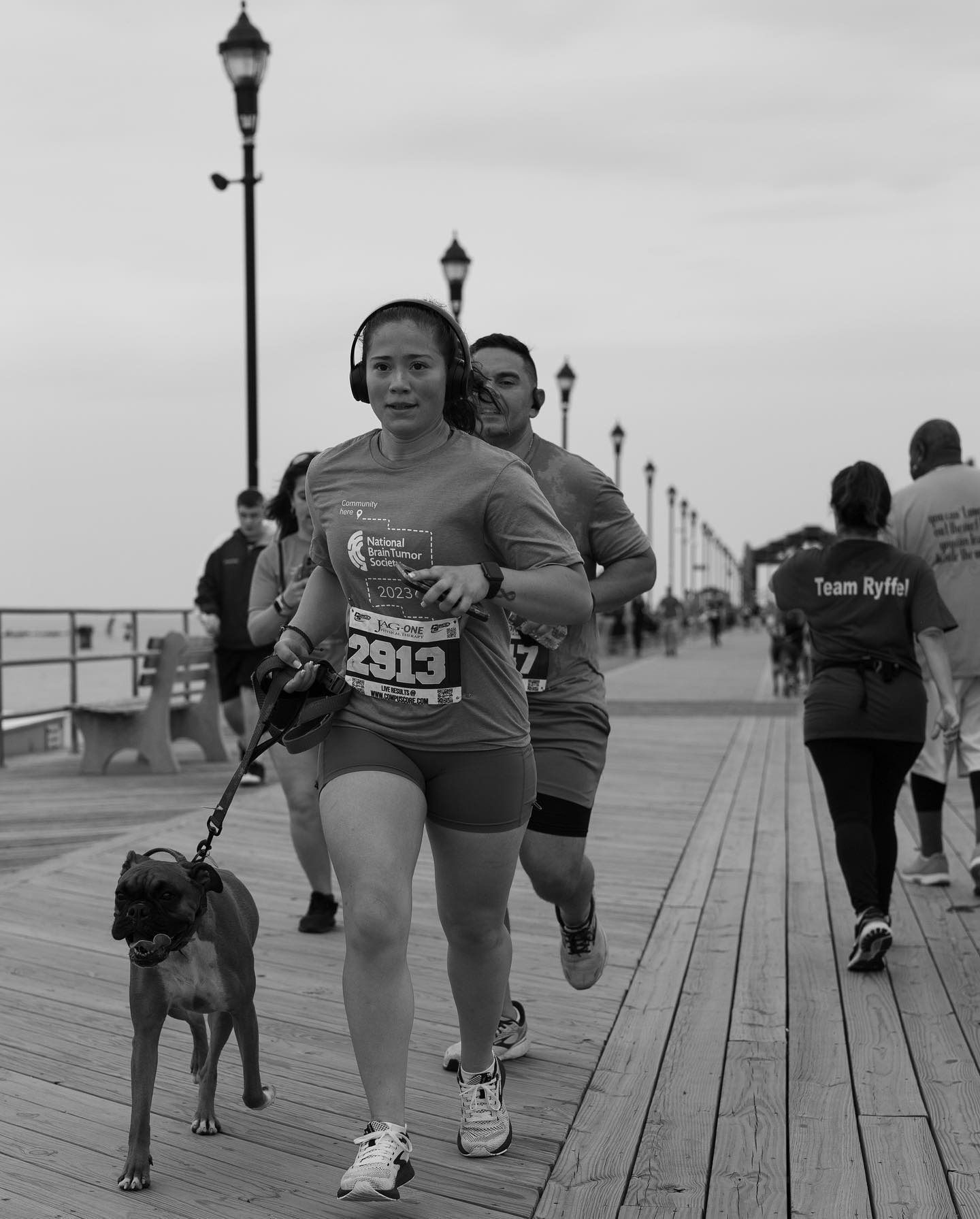 Woman running a race on a boardwalk with a dog on a leash, other runners nearby.