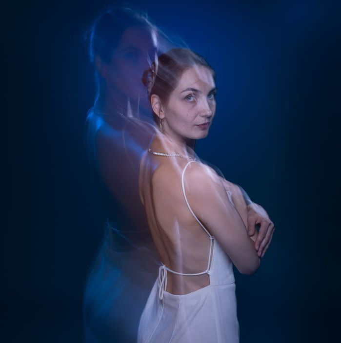 Woman in white dress, arms crossed, with a blurred double exposure effect against a dark blue background.