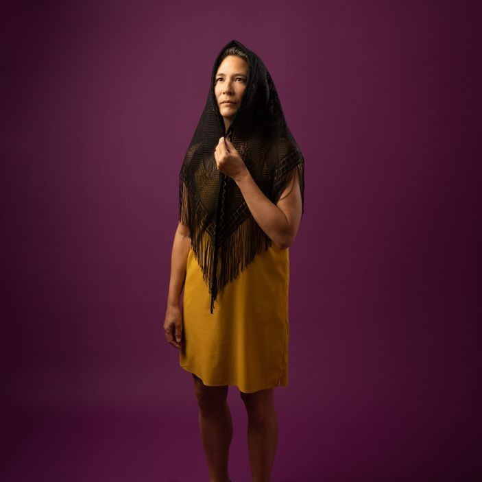 Woman in a yellow dress and black shawl, against a purple background, looking upward.