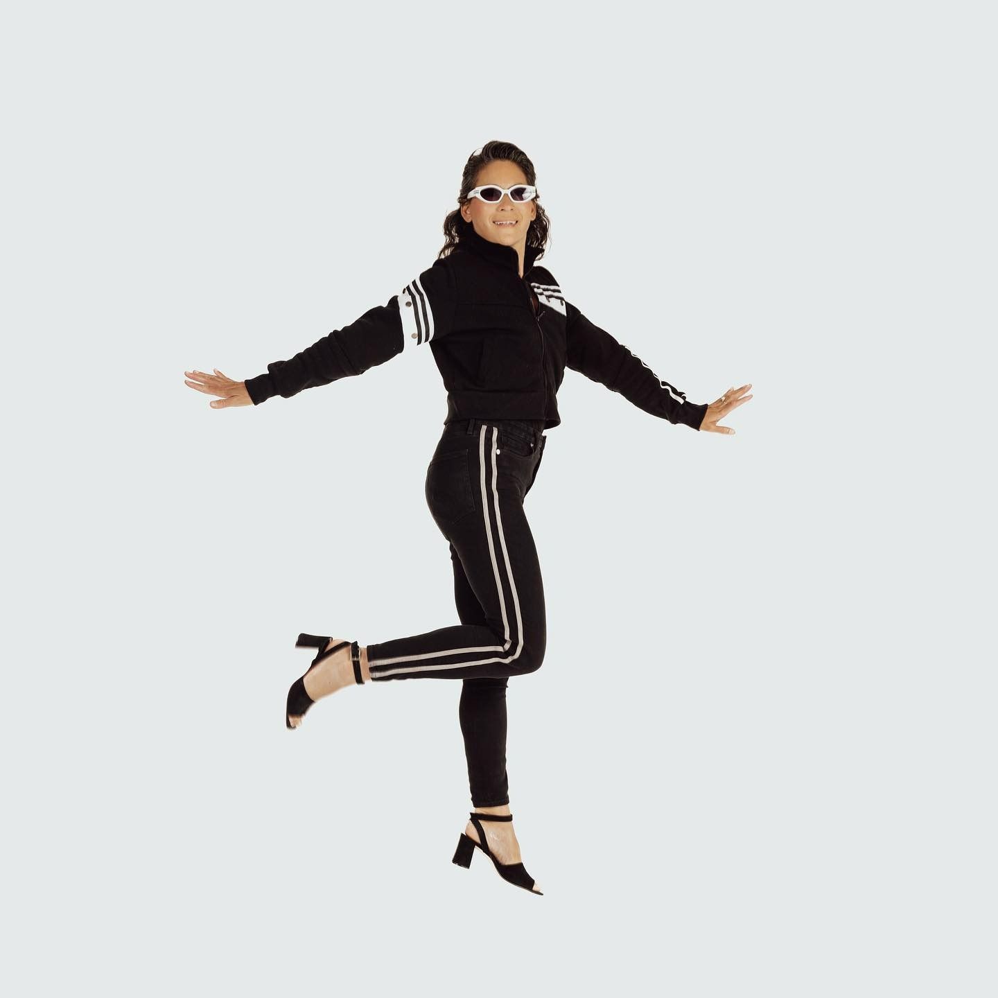 Woman wearing black track suit and sunglasses, balancing on one foot, arms outstretched.