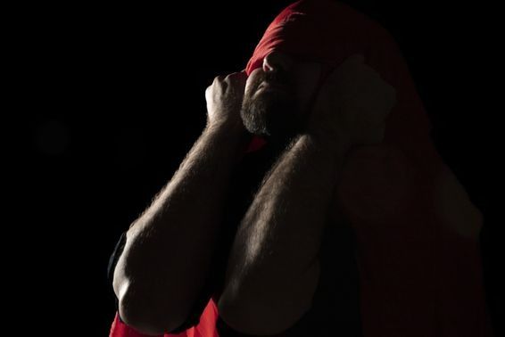 Man in shadows, red cloth partially covering his face, arms raised.