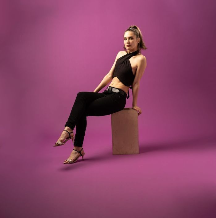 Woman in black outfit sits on a small box against a magenta background.
