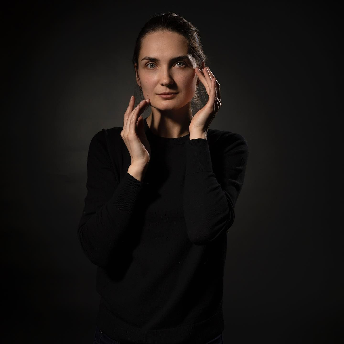 Woman in black sweater, hands near face, looking forward. Dark background.