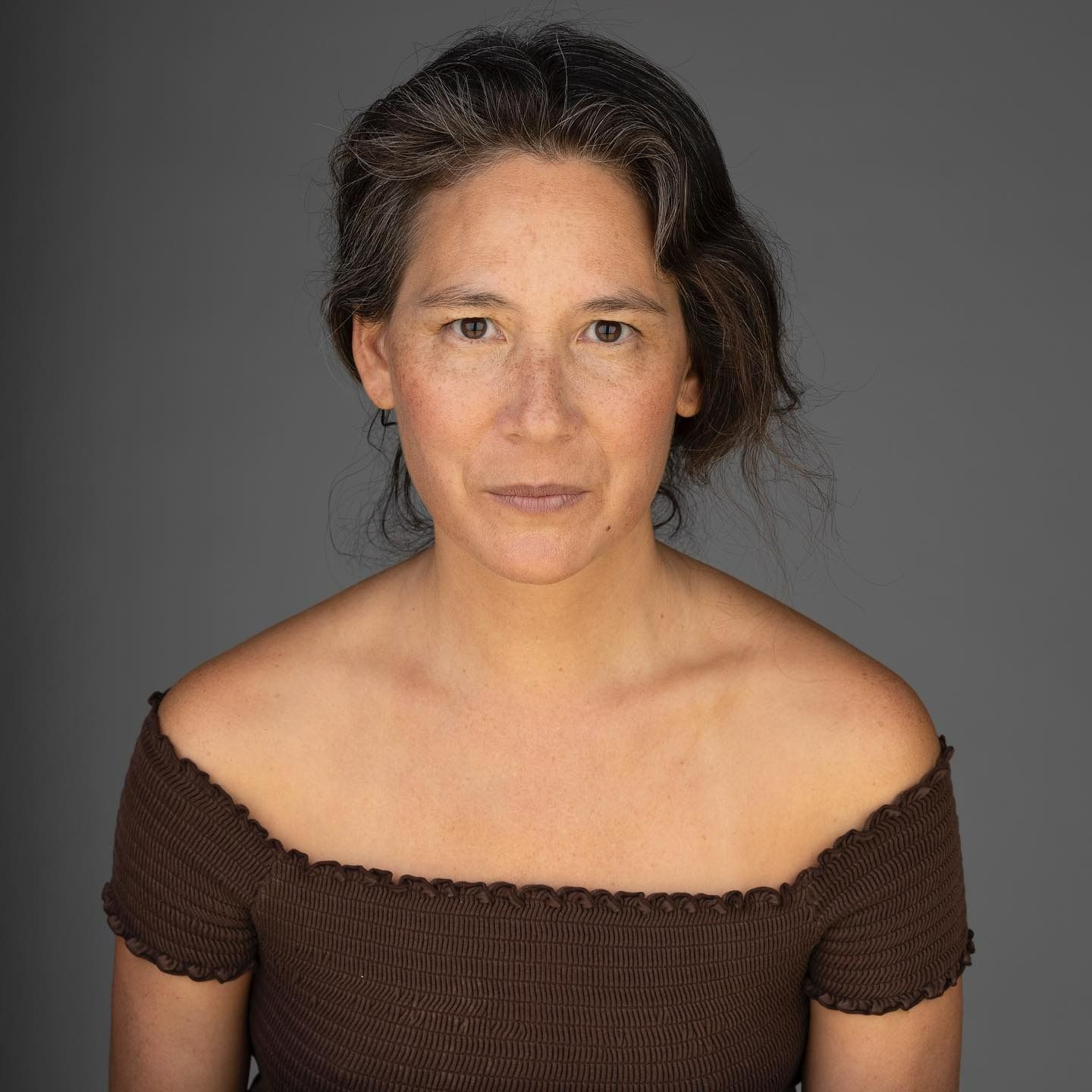 Woman with brown hair and an off-the-shoulder brown shirt against a gray background.