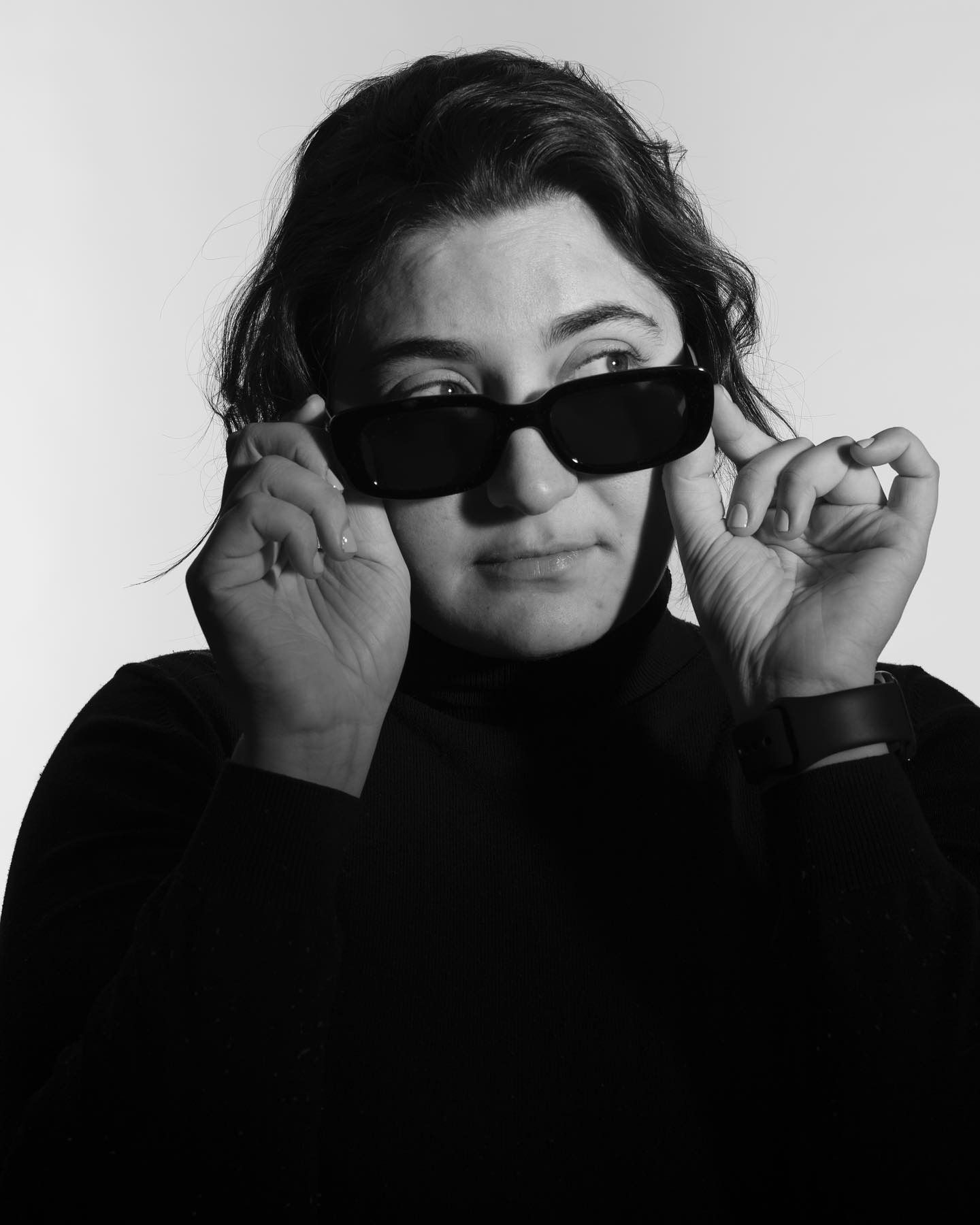 Woman in black turtleneck and sunglasses adjusting them, looking to the side.