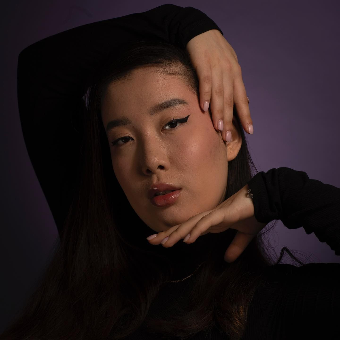 Woman with dark hair, black top, hands framing her face, against a purple backdrop.