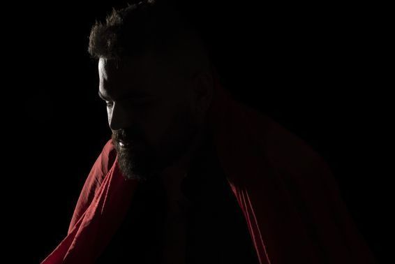 A person with a beard, partially illuminated, wearing red fabric against a black background, looking down.