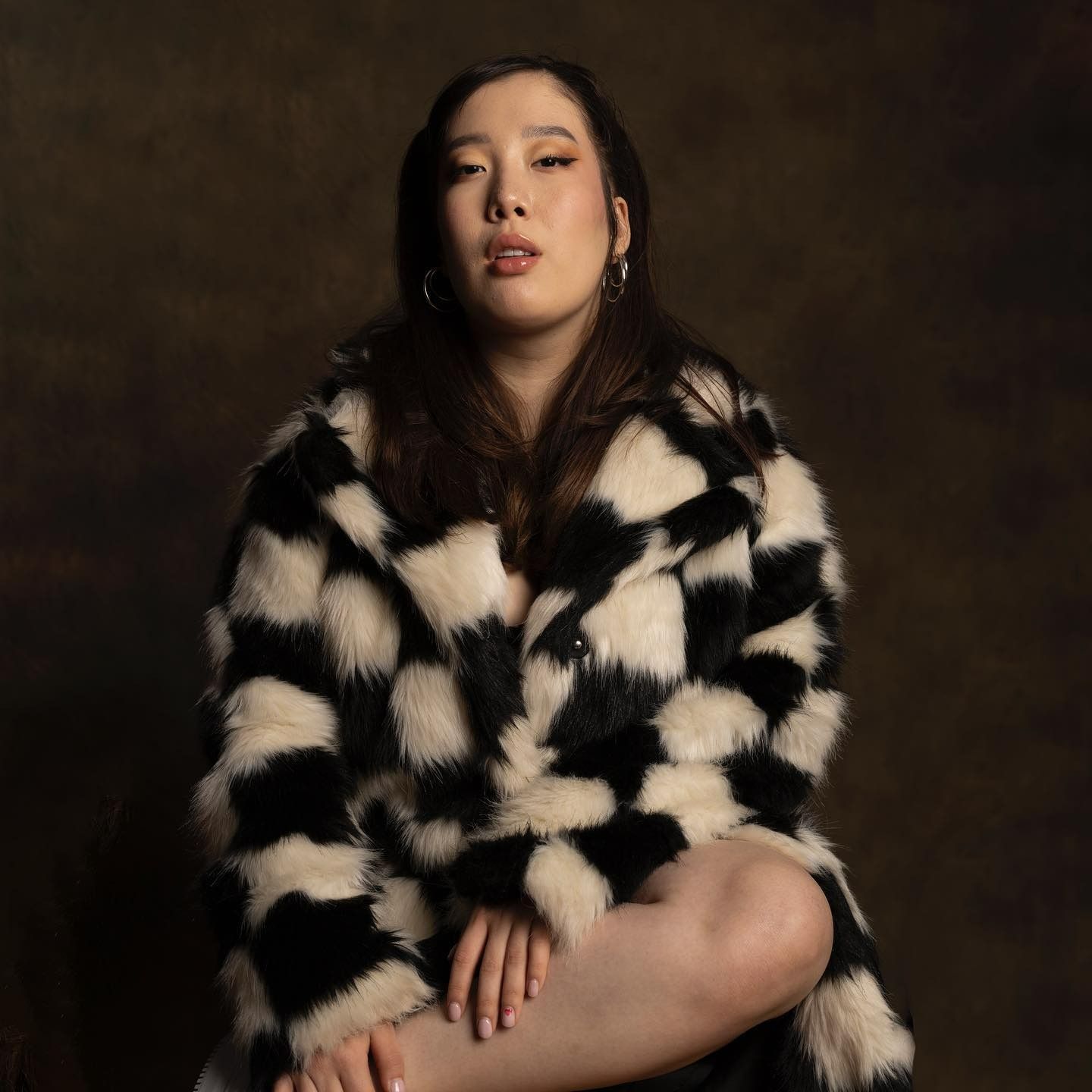 Woman wearing black and white checkered faux fur coat, sitting and looking forward. Brown backdrop.