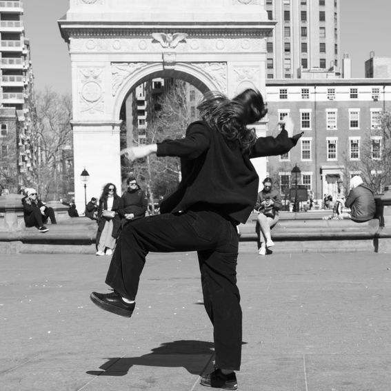 Person dancing in front of Washington Square Arch, arms outstretched, leg raised. Black and white.