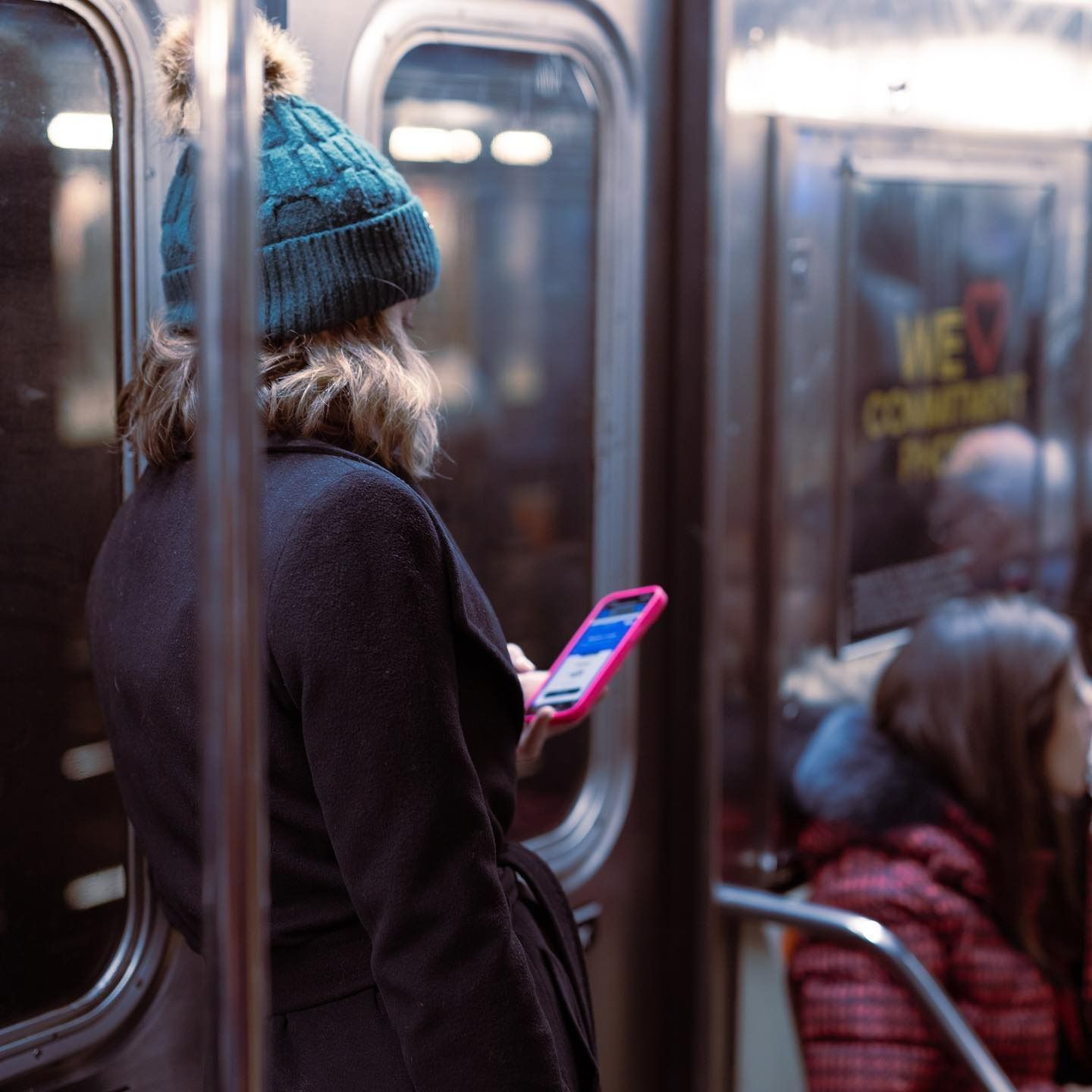 Woman in a teal hat and coat, on a subway, looks at a pink phone.
