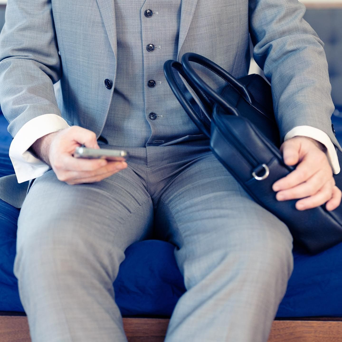 Man in gray suit sits on a blue surface, holding a phone and briefcase.