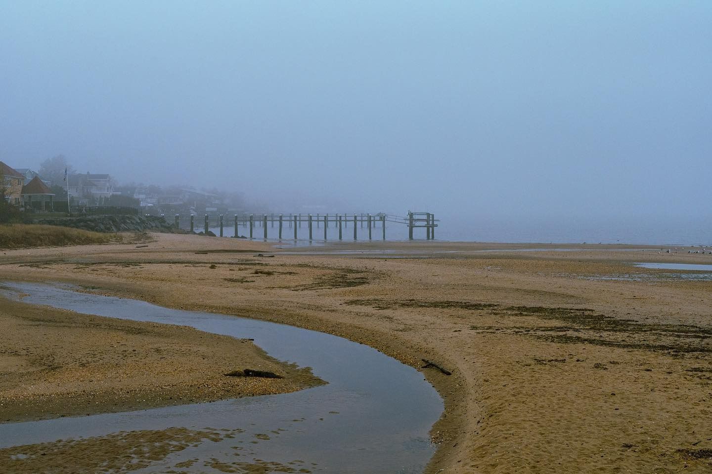 Foggy beach scene with a wooden pier extending into the water. A small stream runs across the sand.