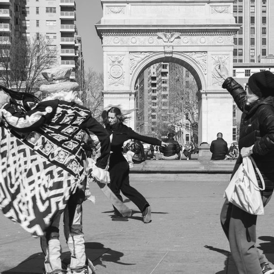 People in a plaza with Washington Square Arch. One person raises arm, others move around. Black and white.