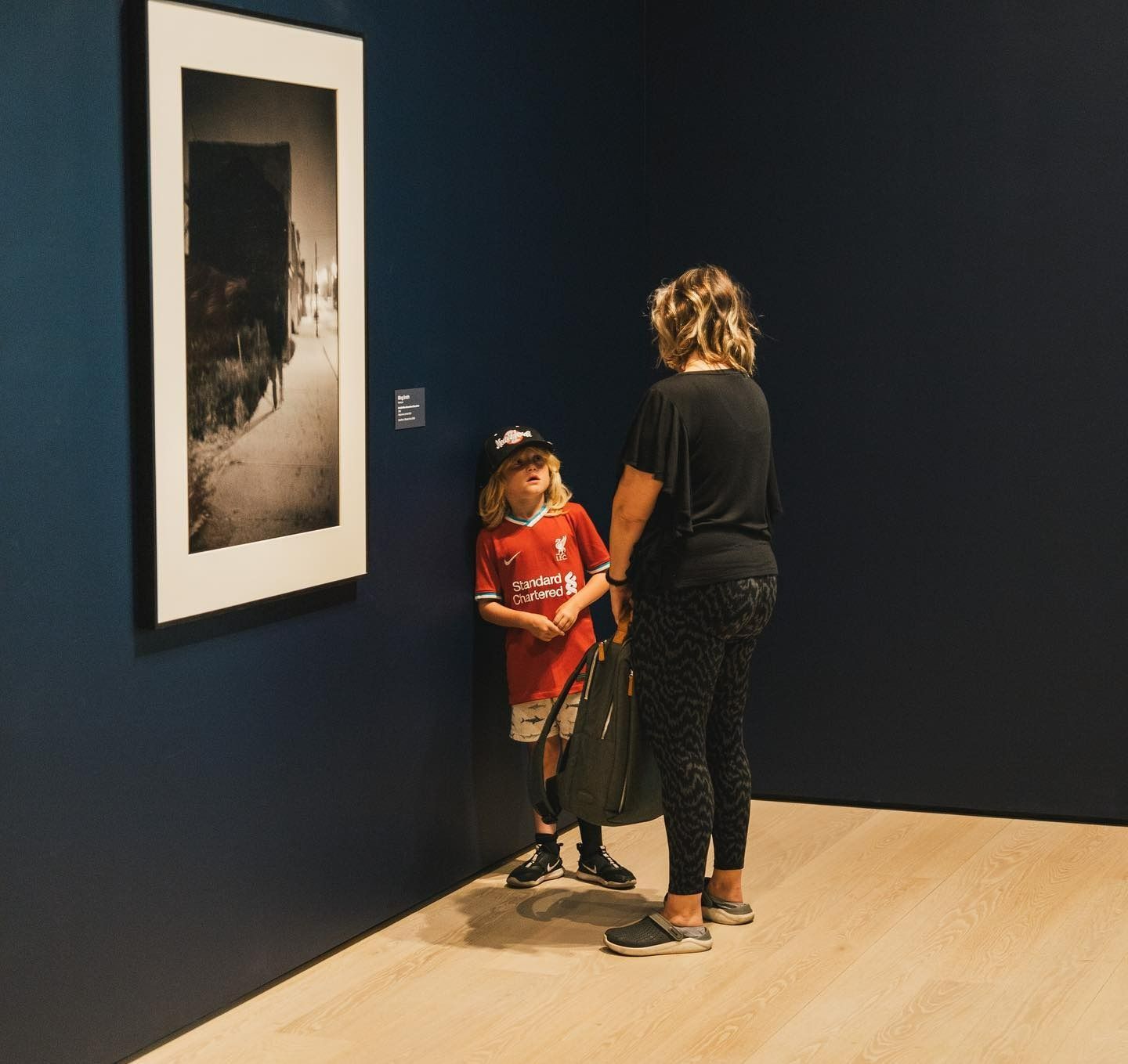 Woman and child viewing artwork in a gallery. Child in red shirt stands near a wall.