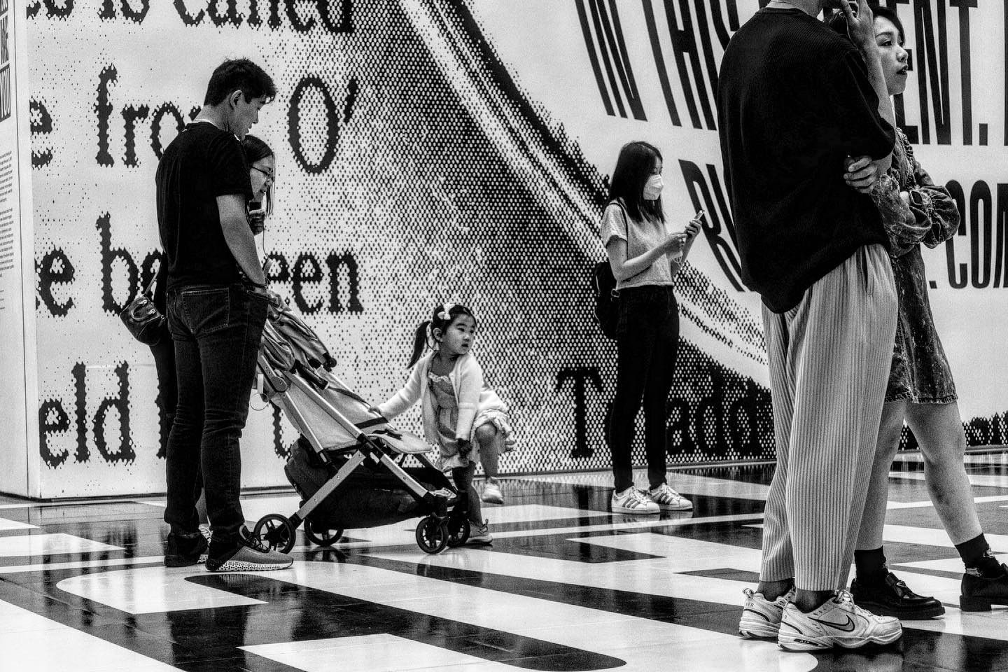 People view a text-covered wall; a child sits in a stroller, others stand and use phones. Black and white.