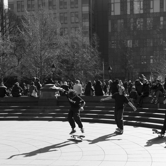 Skateboarders in a sunny public square; one jumps a board, others ride near onlookers and buildings.