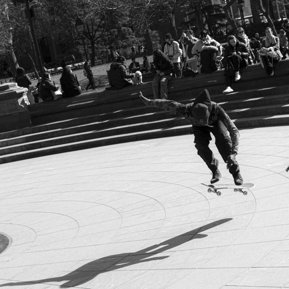Skateboarder in motion, performing a trick on a concrete surface, with onlookers in the background. Black and white.