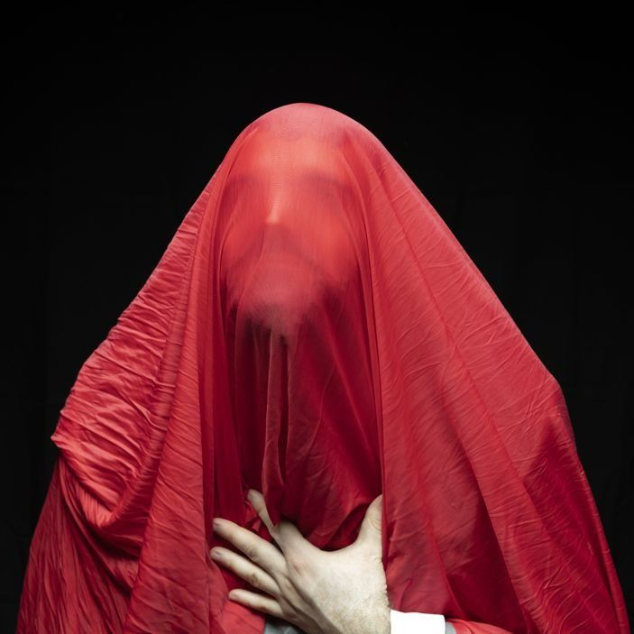 Person covered in red fabric, hand on chest, black background.