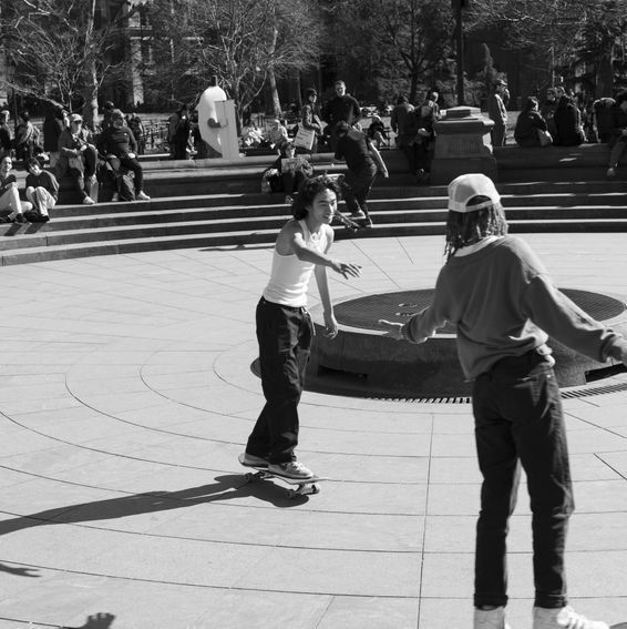 Two people skateboarding in a plaza, one on a board, the other watching. People sitting in the background.