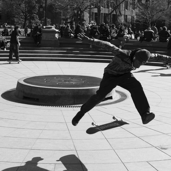 Skateboarder in mid-air above his board, near a fountain. Other people are in the background. Black and white.