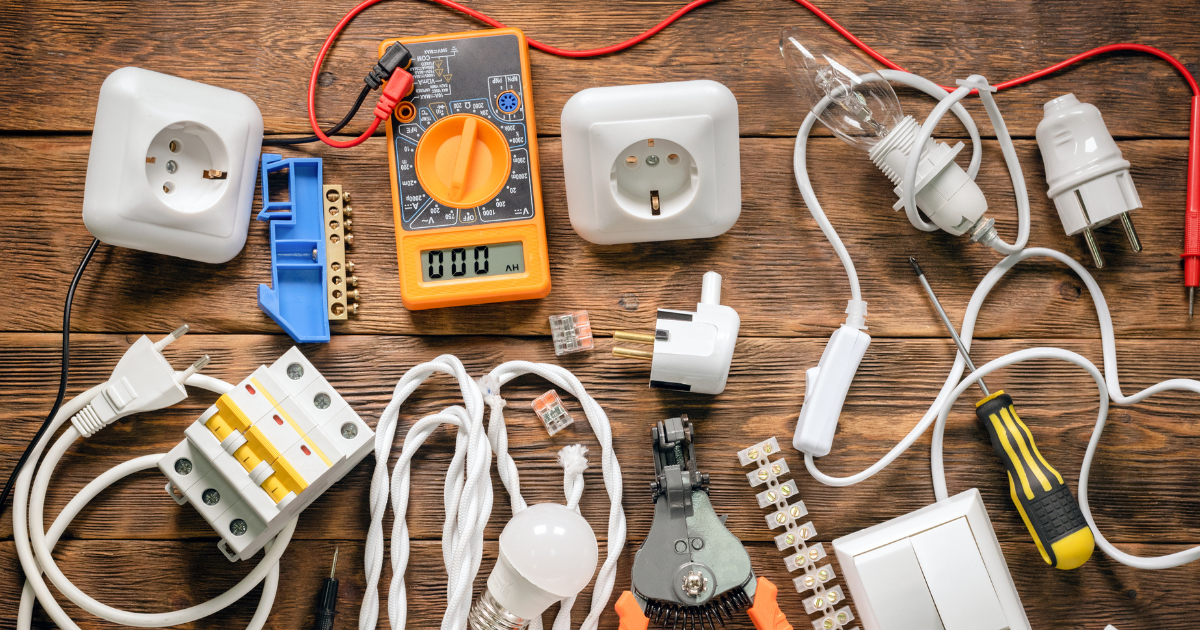 A variety of electrical supplies, including sockets, a multimeter, light bulbs, wires, and tools on a wooden surface.