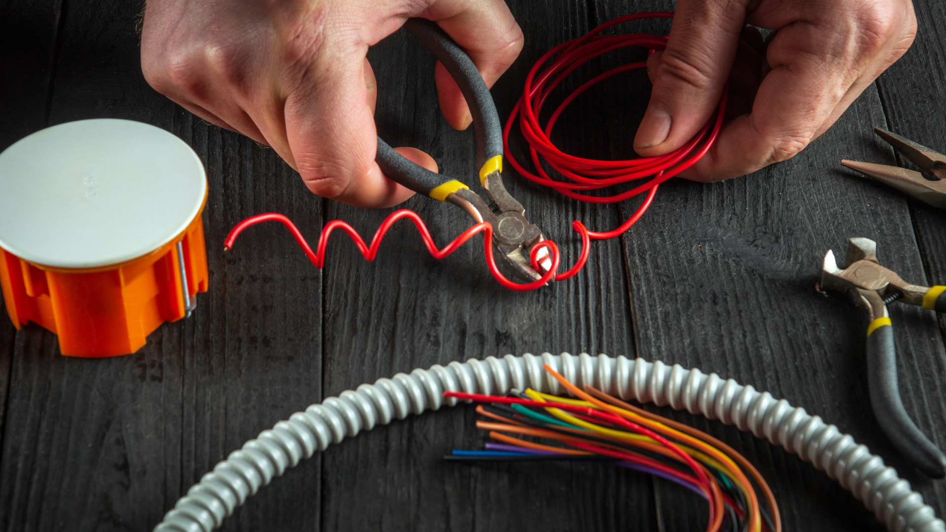 Hands use wire cutters to work with red wire near a plastic electrical box, conduit, and other tools on a wooden surface.