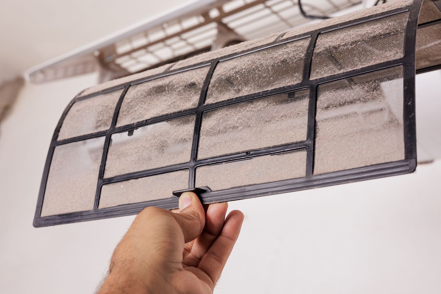 A hand holds a dirty air conditioning filter, with a blurry air conditioner in the background.