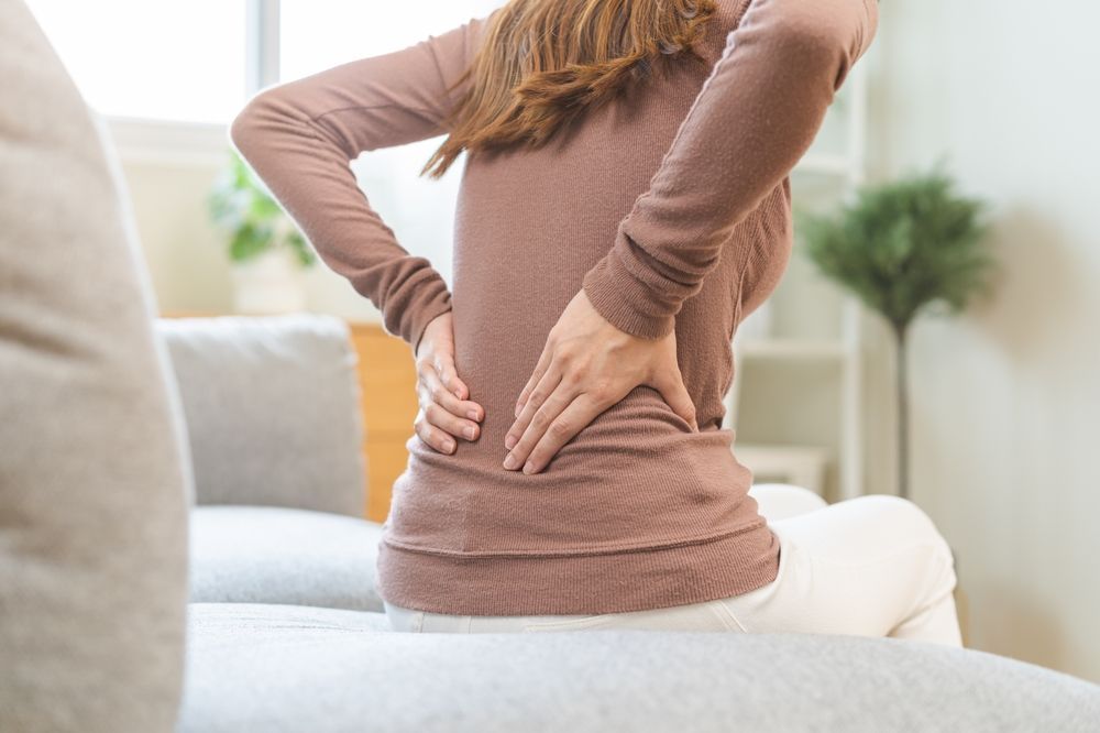 Woman sitting on a couch, holding lower back in pain, indoors.