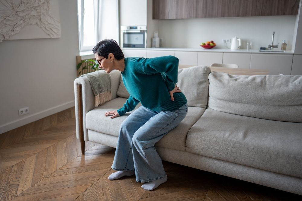 Person clutching side while standing beside a beige sofa in a living room