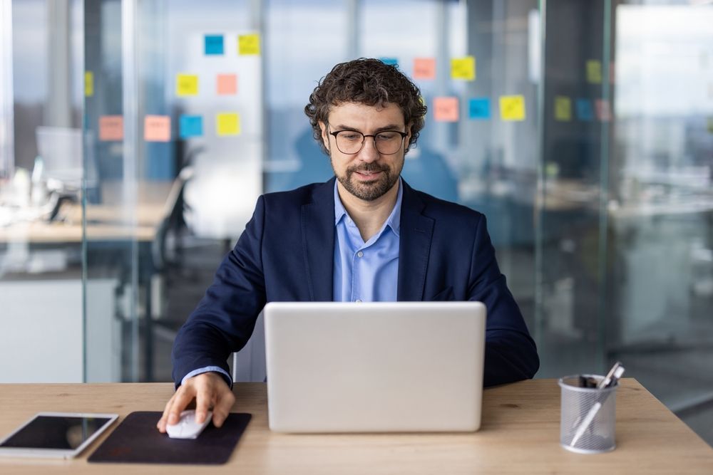 Man working at a laptop in a modern office, with sticky notes on a glass wall behind him.