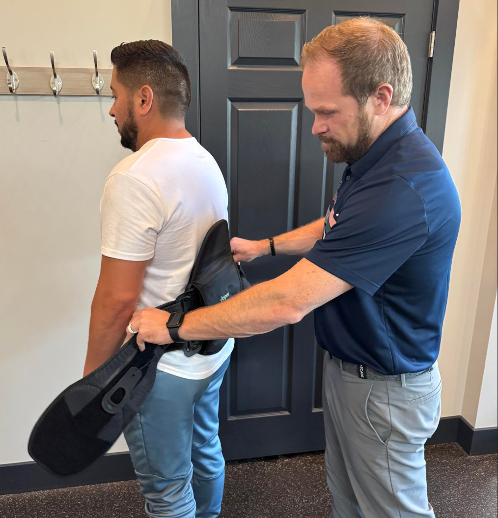 Man receiving lower back brace fitting from another man. Indoors.