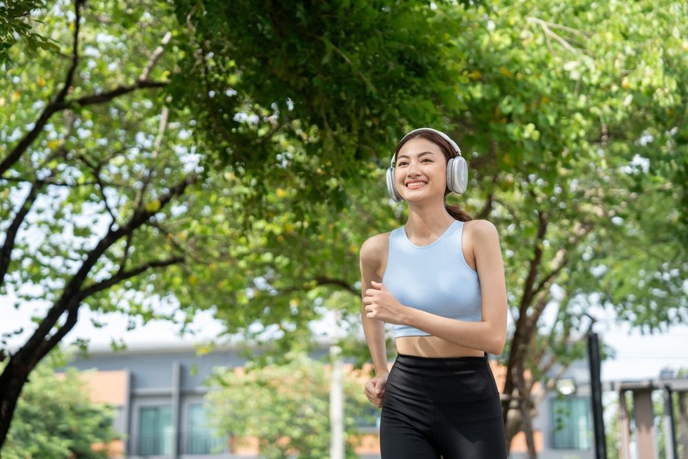 Woman jogging outdoors, wearing headphones and smiling, trees in background.