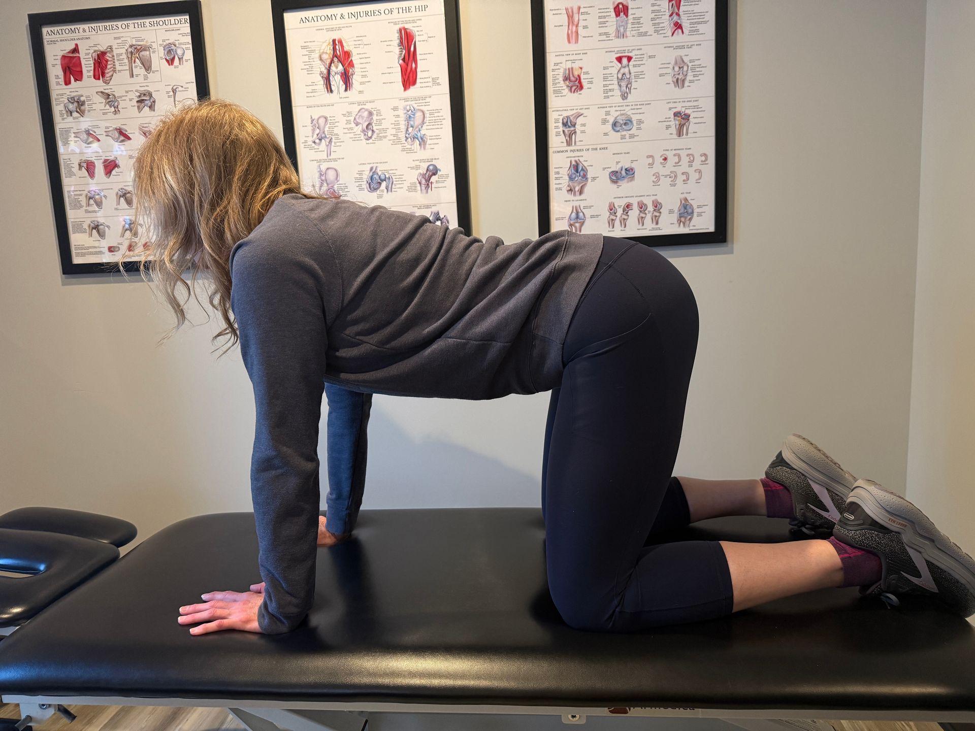 Woman on hands and knees, doing an exercise on a black table, with anatomical charts on the wall.