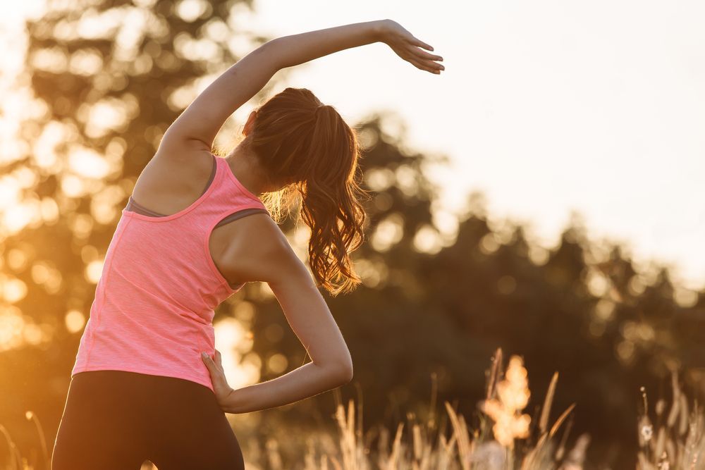 Woman in pink tank top stretching outdoors at sunset.