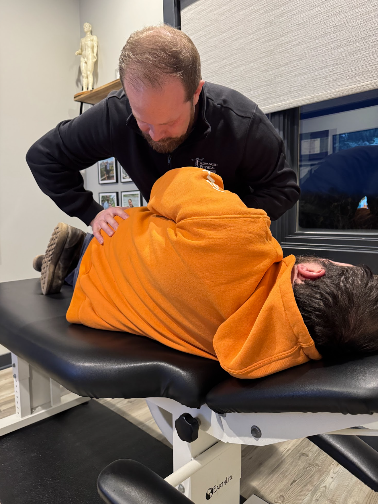 Man receiving chiropractic adjustment lying on a table, orange towel, healthcare provider nearby.