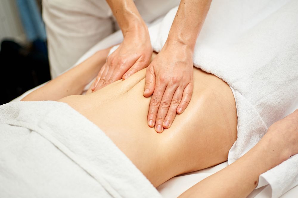Hands massaging a person's abdomen, covered by a white towel, in a massage setting.