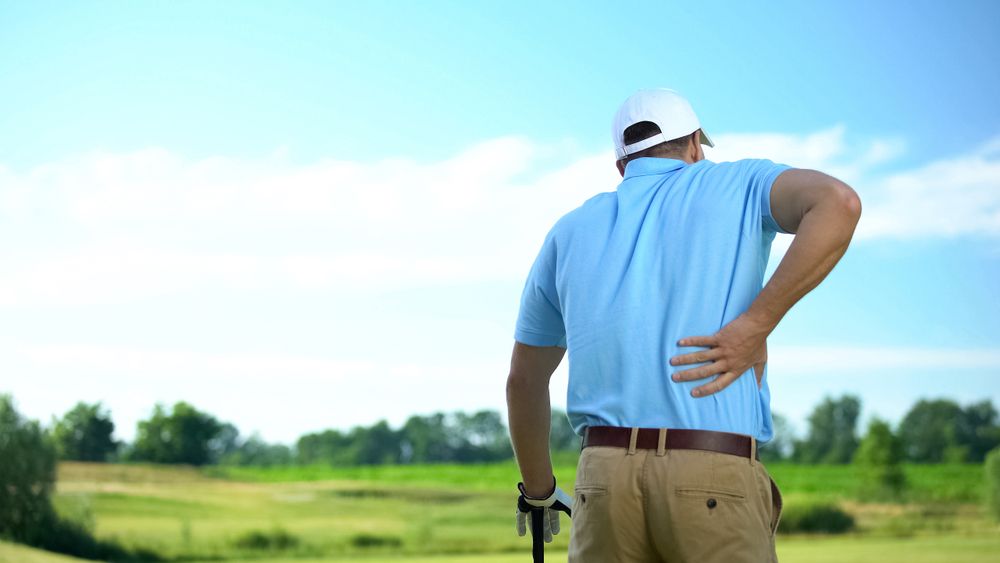 Golfer in a blue shirt and white cap stands on a green course, holding his back