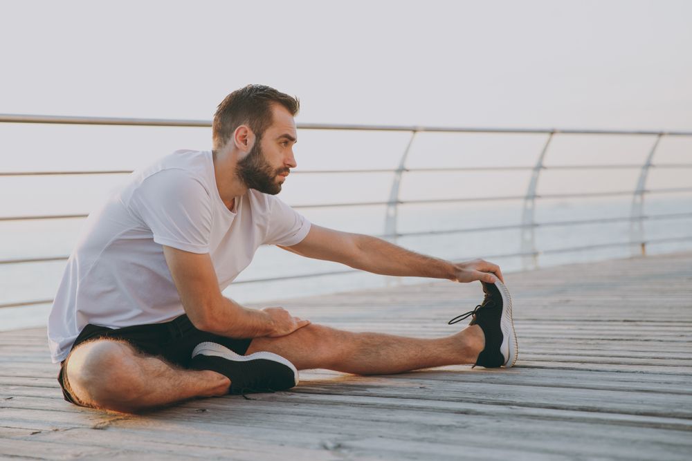 A man is sitting on a wooden pier stretching his legs.