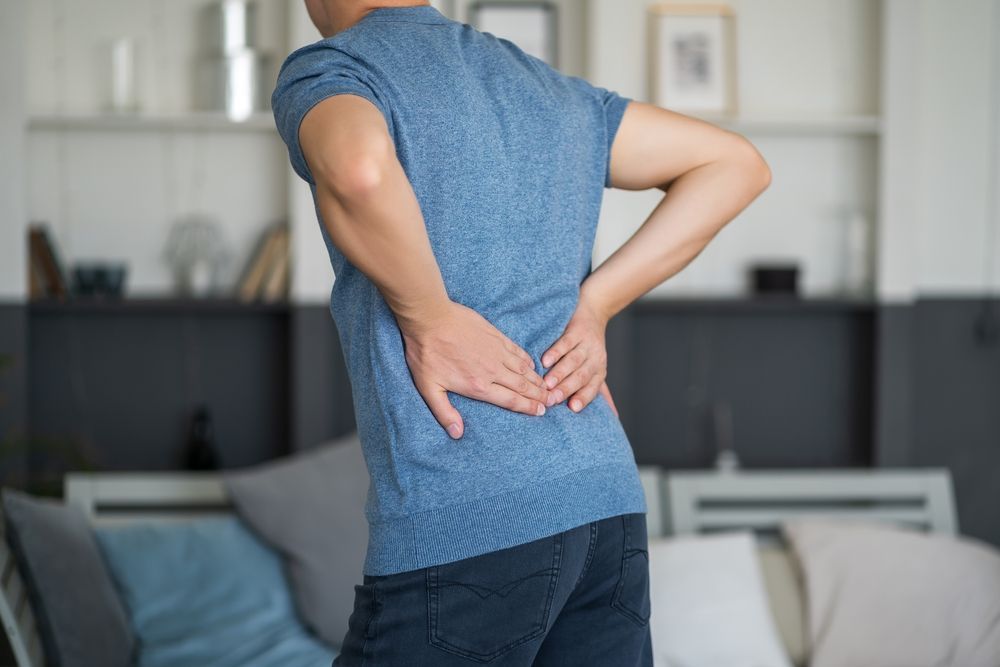 A man is holding his back in pain while standing in a living room.