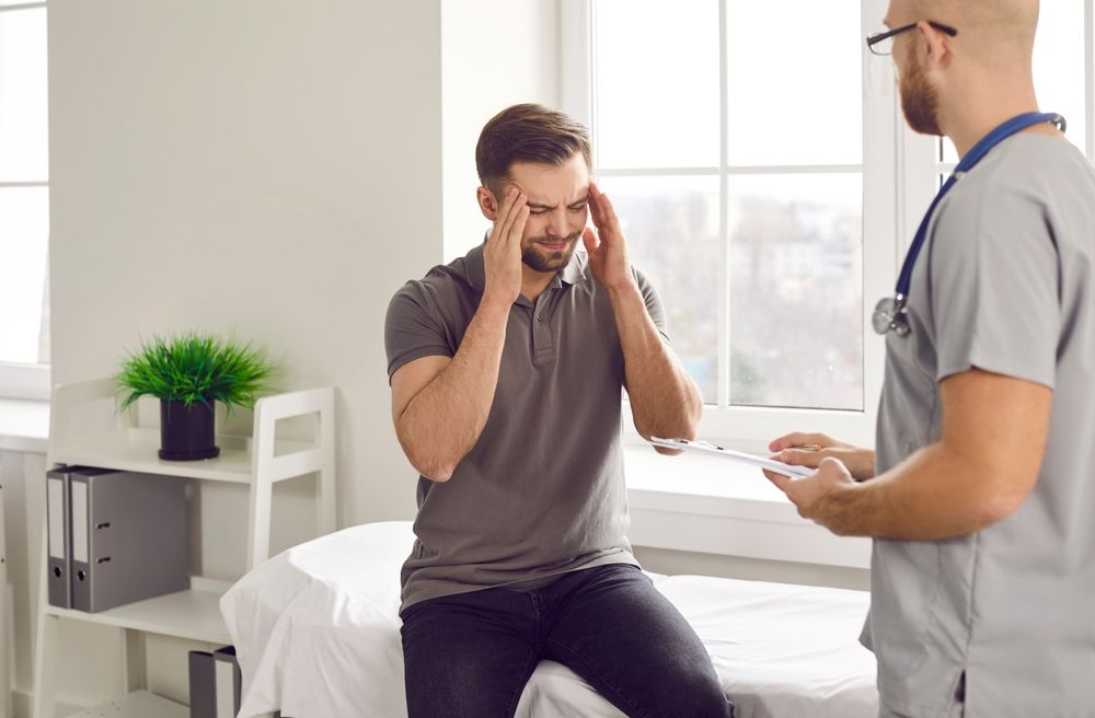 a man with a migraine is sitting on a bed talking to a chiropractor.