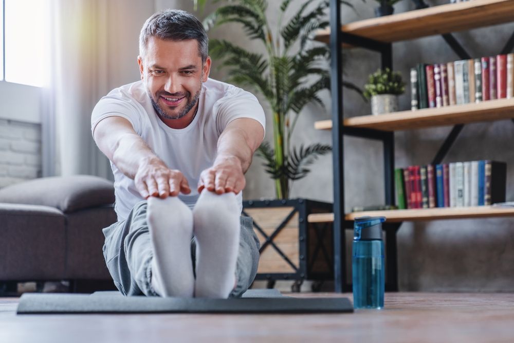 Man in white shirt stretching on a yoga mat indoors, smiling.
