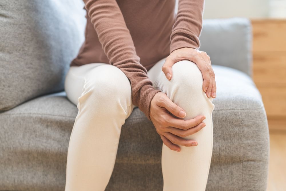 A person sitting on a gray couch, holding their knee with both hands as if in pain.